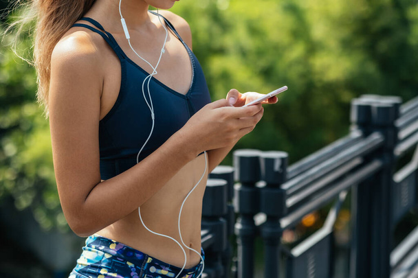 Unrecognizable fitness woman holding mobile phone and listening music on earphones in outside .Fit woman in sportswear using mobile phone to listen music and to track fitness progress on app after jogging in city - Photo, Image