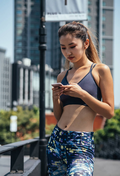 Portrait of woman in sportswear using mobile phone in city. Sports woman resting and checking results on smartphone app after jogging in the city - Fotografie, Obrázek