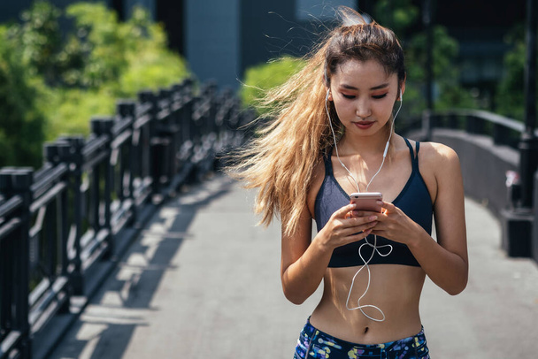 Portrait of fit woman in sportswear using phone to listen music before workout outdoors. Beautiful young sporty woman listening music on smart phone on bridge in the city - Foto, Bild