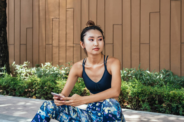 Attractive woman resting after running. She is sitting on sidewalk and using smartphone. Young woman resting and checking results on smartphone app after jogging in the city - Foto, immagini