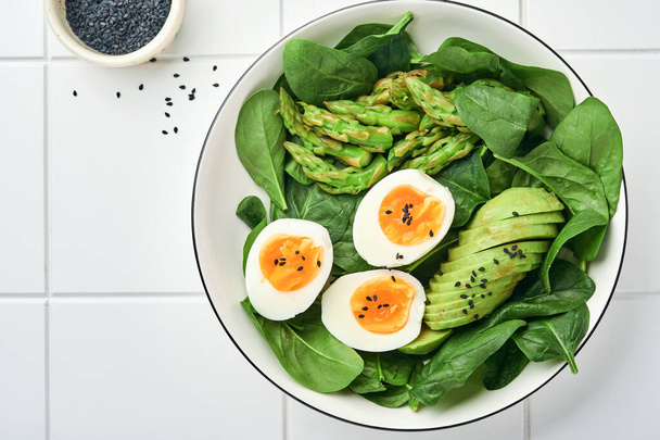 Fresh vegetable salad with avocado, asparagus, crumpled eggs with black sesame seeds and spinach on plate on light slate, stone or concrete background. Balanced lunch in bowl. Top view. Mock up. - Foto, Bild