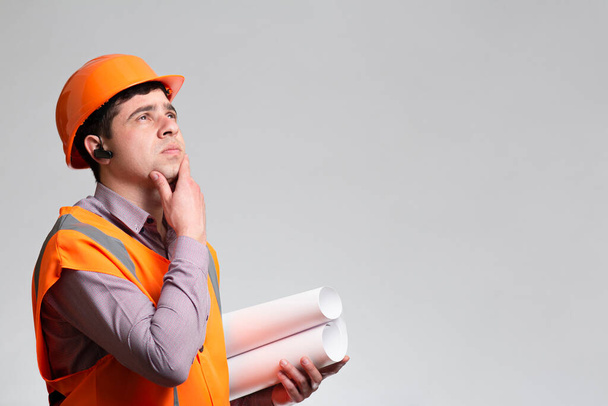 Young constructor in hard hat and reflective vest thinking with hand on chin on grey studio background, foreman examines building project - Foto, Imagem