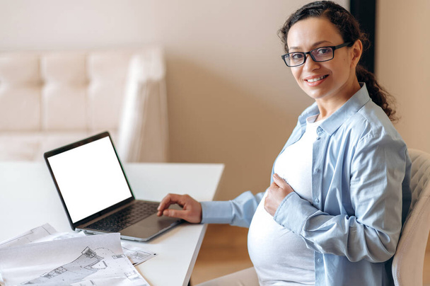 A pregnant woman, designer or freelancer, sits at a work desk, gently hugs her tummy with her hand, looks and smiles into camera, on the table a laptop with blank white screen and a working documents - Foto, afbeelding
