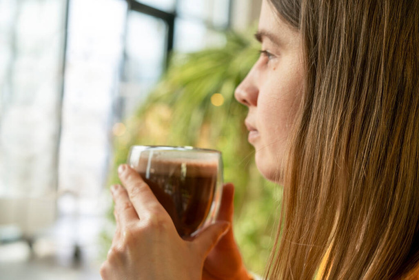 Genç, beyaz bir kadın elinde bir bardak kahve ya da kakao tutarak pencereden dışarı bakıyor yeşil ev bitkilerinin arka planına bakıyor, kahve saati, sıcak içecekler, sıcak çikolata... - Fotoğraf, Görsel