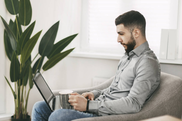 Beau jeune homme d'apparence orientale travaille à la maison, assis sur une chaise avec un ordinateur portable - Photo, image