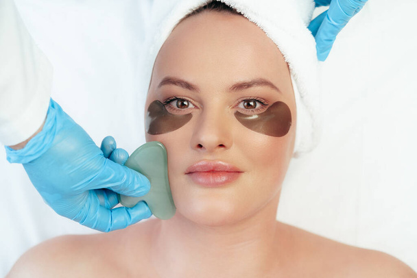 Beautiful woman having guasha scraper face massage done by cosmetologist - Фото, зображення