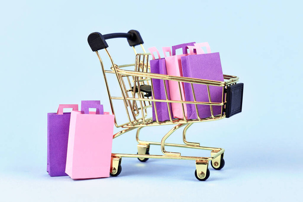 Golden shopping cart filled with pink and purple paper shopping bags on blue background - 写真・画像
