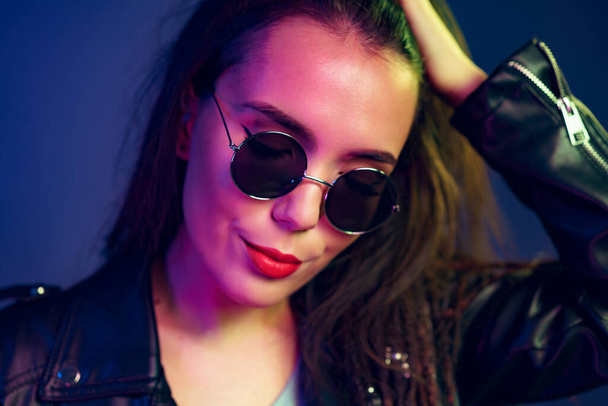 Asian girl in sunglasses and black leather jacket posing against dark blue background - Фото, зображення