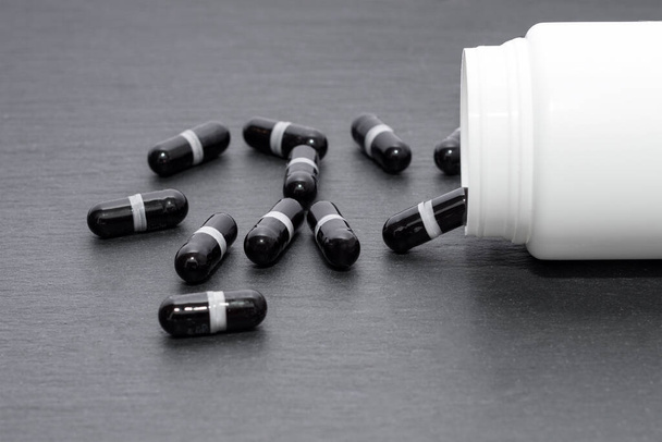 Czarne kapsułki suplement są wylewane z białej butelki na tablicy. Temat medyczny, koncepcja opieki zdrowotnej - Zdjęcie, obraz