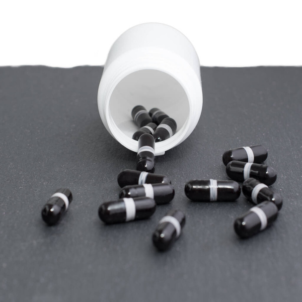 Черные капсулы добавки выливаются из белой бутылки на доску. Тема медицины, концепция здравоохранения - Фото, изображение