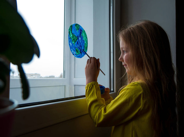 Evdeki pencereye gezegeni boyayan sevimli küçük kız. 22 Nisan Dünya Günü 'nüz kutlu olsun. Yaratıcı aile eğlencesi yeni gerçekliği kilitliyor. Çevrenin bilinçli tüketim kavramının korunması. - Fotoğraf, Görsel