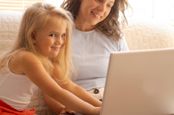 Onnellinen pieni esikoulutyttö yhdessä äidin kanssa kannettavan tietokoneen avulla, istuu sohvalla. Sarjakuvan etsiminen, videopuhelun tekeminen isovanhempien tai isän kanssa, rentouttava hauskanpito, ostokset verkossa, selailu - Valokuva, kuva