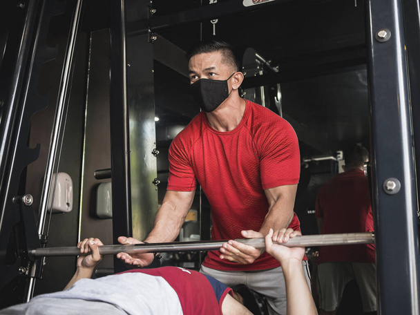 Ένας γυμναστής με μάσκα προσώπου εντοπίζει τον πελάτη του να κάνει πάτημα πάγκου με εναλλασσόμενη λαβή.. - Φωτογραφία, εικόνα