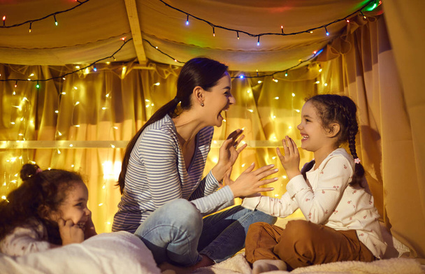 Happy mother playing with adorable cheerful children in home tent at evening - Foto, Bild