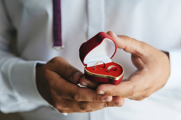 Pair of gold wedding rings in red heart-shaped box in hands of groom on wedding day horizontal shot - Фото, изображение