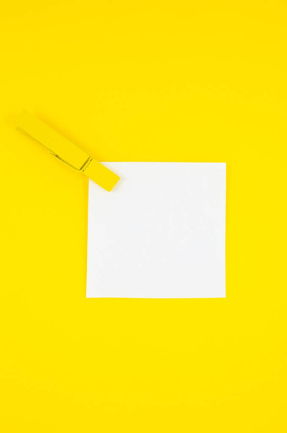 Λευκό αυτοκόλλητο για να κάνετε λίστα με κίτρινα ξύλινα μανταλάκια. Κοντινό πλάνο του σημειώματος υπενθύμισης χαρτί στο κίτρινο φόντο. Αντιγραφή χώρου. Μινιμαλισμός, πρωτότυπη και δημιουργική φωτογραφία. - Φωτογραφία, εικόνα