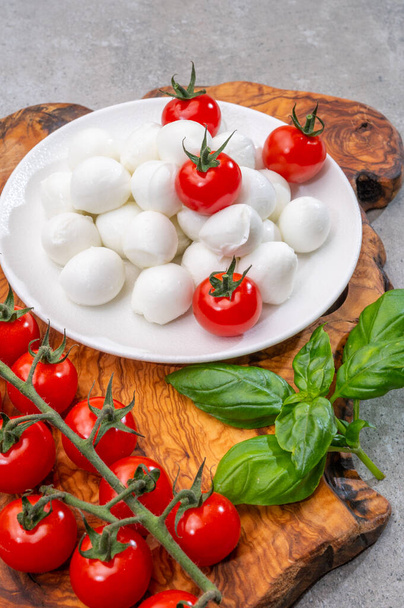 Italialainen tricolore, pienet pallot tuoretta valkoista pehmeää italialaista mozzarella-juustoa, kypsiä punaisia kirsikkatomaatteja ja tuoretta vihreää basilikaa, valmis caprese-salaatin valmistukseen, lähikuva - Valokuva, kuva