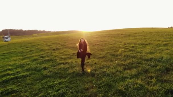 Vista aérea de una joven de pelo largo con una gabardina negra corriendo sobre un prado verde sobre un fondo al atardecer - Imágenes, Vídeo