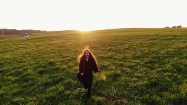 Siyah trençkotlu genç bir kızın gün batımında dalgalanan saçlarıyla yeşil bir çayırda koşarkenki görüntüsü. - Video, Çekim