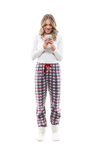 Glückliche junge hübsche Frau im bequemen Pyjama beim Internetsurfen auf dem Smartphone. Volle Körperlänge isoliert auf weißem Hintergrund. - Foto, Bild