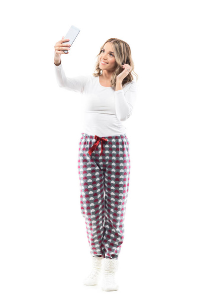 Stunning beauty woman in pajamas taking selfie with smart phone touching hair. Full body length isolated on white background. - Photo, Image