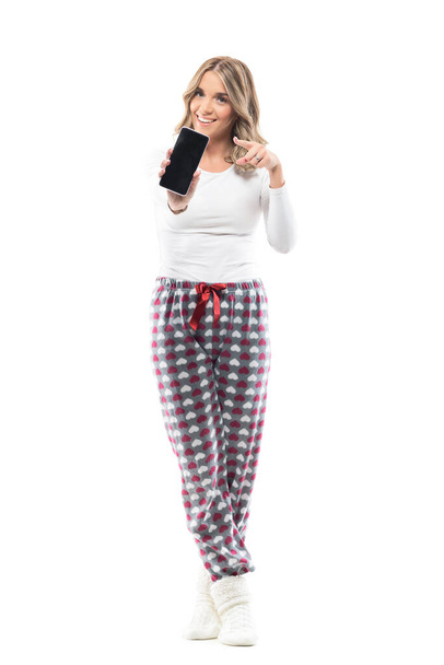 Hezká žena v pyžamu, držící a ukazující černou obrazovku mobilu ukazující prstem a reklamu. Plná délka těla izolované na bílém pozadí. - Fotografie, Obrázek