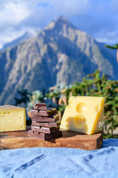 Sabrosos quesos suizos y chocolate negro puro, emmental, gruyere, appenzeller servido al aire libre con picos de las montañas alpinas en verano en el fondo - Foto, Imagen