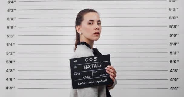 Mugshot of arrested young woman turning head and looking to camera while standing aside. Crop view of pretty female criminal with ponytail holding sign for photo in police department. - Footage, Video