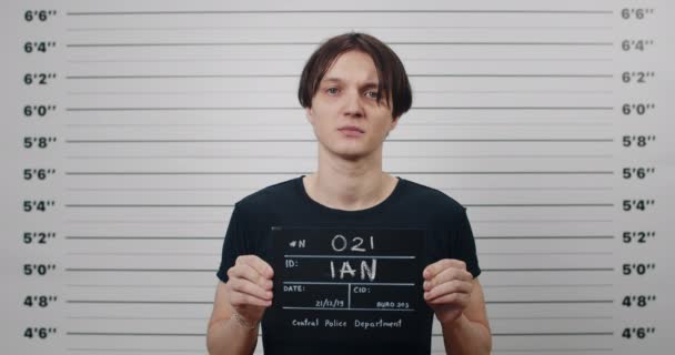 Portrait of young male criminal person holding sign for photo in police department. Crop view of serious guy posing, raising head and looking to camera in front of lineup metric wall. - Filmati, video