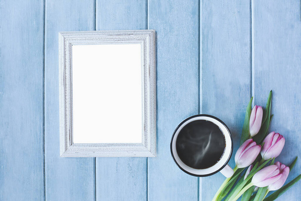 Vertical empty picture frame with steaming hot cup of black coffee and pink spring tulip flowers over a rustic blue wooden table shot from top view. - Fotografie, Obrázek