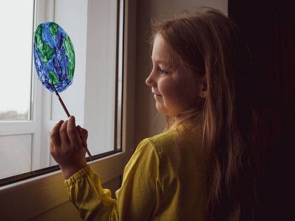 かわいい女の子が家の窓に惑星を描く。ハッピーアースデイ4月22日挨拶メッセージ。創造的な家族のレジャーは新しい現実をロックダウン。生態系の保全環境に配慮した消費コンセプト. - 写真・画像