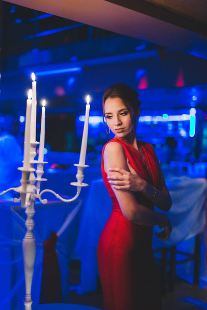 A pretty Caucasian lady wearing red evening dress and posing with white burning candles on foreground - Photo, Image