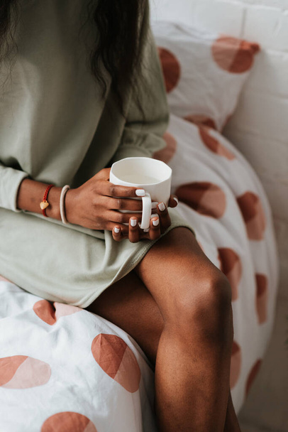 A closeup of a young African female holding a white ceramic mug while sitting on the bed - Fotografie, Obrázek