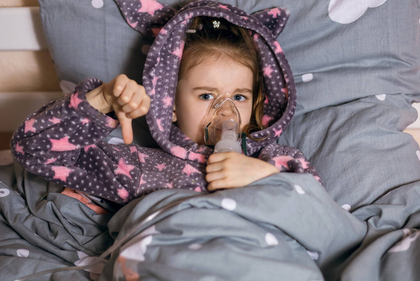 A cute little girl in a hooded robe got sick and is lying on the bed with a stationary inhaler. A severe cough is treated with steam at home. The child shows a gesture with a thumb down - Фото, изображение