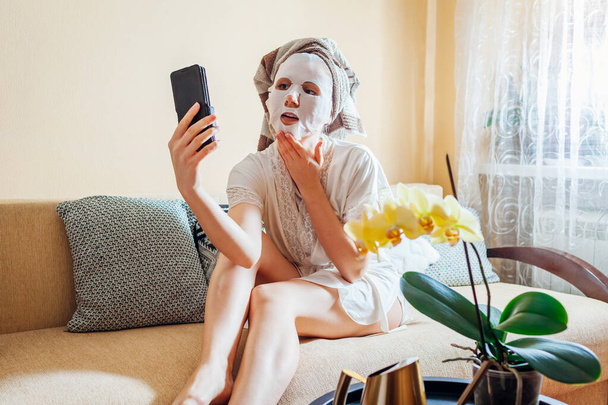 顔のシートマスクをした女性は、自撮りしたスマートフォンを使って自宅でリラックスして頭の上にタオルを着用しました - 写真・画像