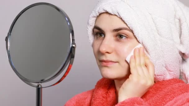 Una mujer joven con una bata rosa se aplica tónico en la cara y el cuello con una almohadilla de algodón. tonificación facial de la piel, prevención de arrugas en las mujeres - Imágenes, Vídeo