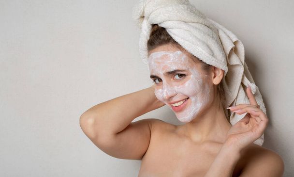 Mujer joven coqueta con toalla de baño en la cabeza usando mascarilla hidratante en su cara. Cuidado personal en el hogar. Espacio para texto - Foto, Imagen