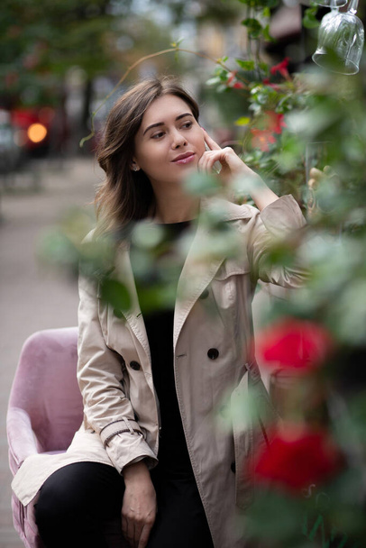 Flirty young woman wears beige coat sitting at the street restaurant in the morning. Outside decoration with green and flowers - Photo, Image