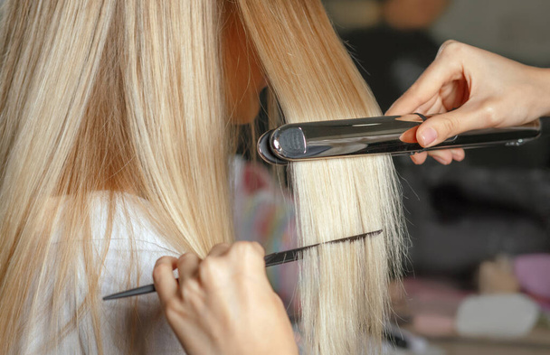 Крупный план рук парикмахера выпрямляет волосы утюгом и плоской расческой - Фото, изображение