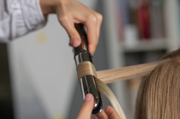 Close-up shot van een kapper het maken van krullen met styling ijzer naar een blonde vrouw bij de kapper  - Foto, afbeelding