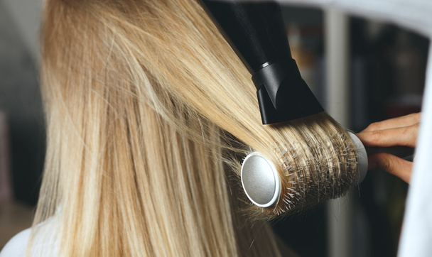 Friseur trocknet im Schönheitssalon die Haare des Kunden mit Bürstenkamm und Föhn - Foto, Bild