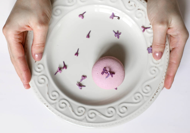 vista recortada de la mujer sosteniendo plato con rosa delicioso macarrón francés o macaron con flores de color lila sobre fondo blanco. - Foto, imagen