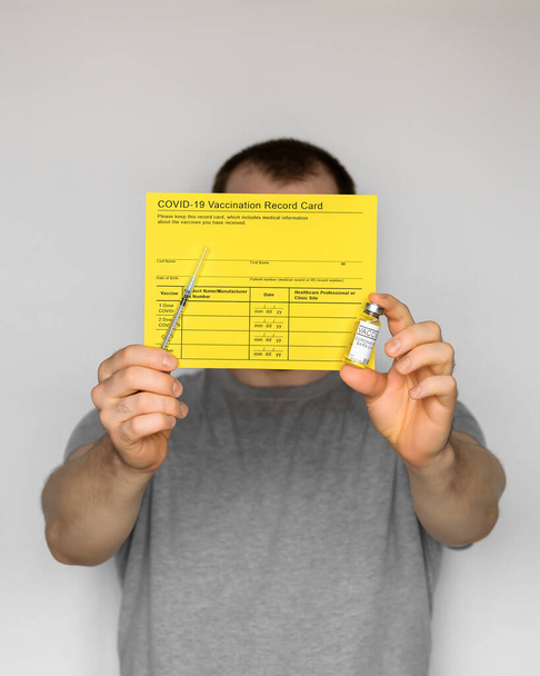 Взрослый мужчина в футболке с жёлтой карточкой для вакцинации, шприцами и пробирками с вакциной COVID-19 для двухступенчатой инъекции. Серый фон - Фото, изображение