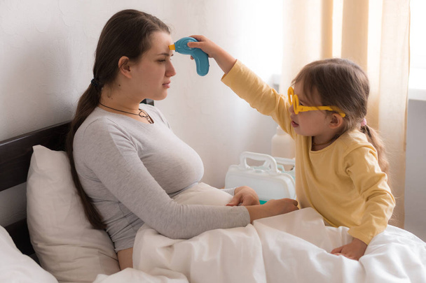 little girl wearing uniform playing doctor, nurse with young mum, nanny in bedroom, checking mother throat, measures temperature, take care, family spend leisure time at home together role play game - Photo, Image