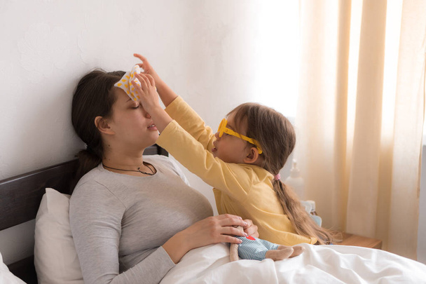 little girl wearing uniform playing doctor, nurse with young mum, nanny in bedroom, checking mother throat, measures temperature, take care, family spend leisure time at home together role play game - Φωτογραφία, εικόνα