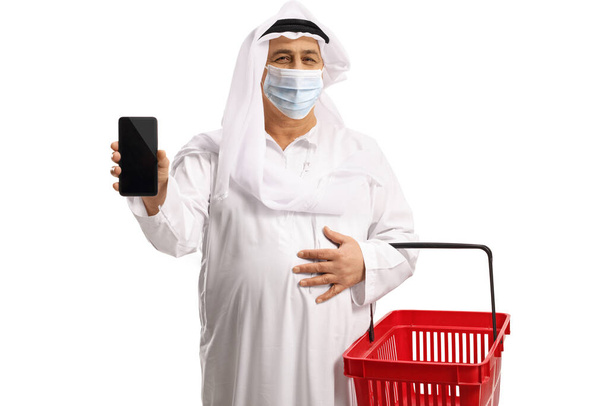 Arab man wearing a protecitive face mask and holding a shopping basket and a smartphone isolated on white background - Foto, afbeelding