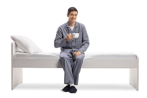 Young man in pajamas sitting on a bed and holding a cup of tea isolated on white background - Foto, imagen