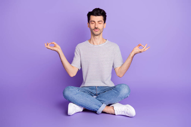 Photo of peaceful guy sit lotus pose meditate wear striped white t-shirt jeans sneakers isolated violet background - Photo, Image
