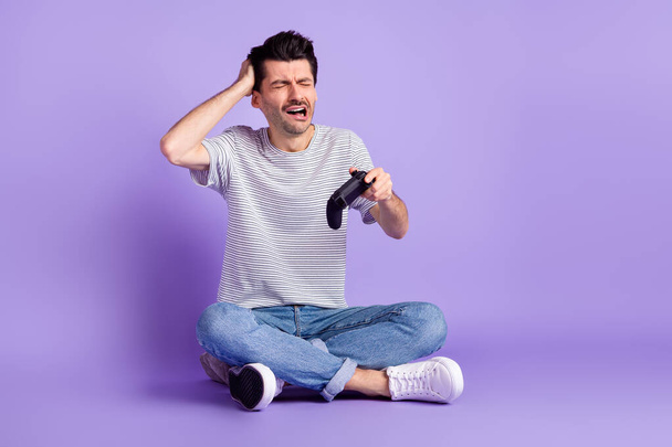 Photo of guy hold gamepad close eyes cry hand head wear t-shirt jeans footwear isolated violet background - Foto, imagen