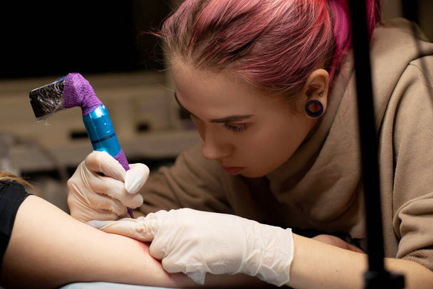 Чарівна дівчина з рожевим волоссям та татуюваннями набиває татуювання на руці жінки в татуювальному салоні, творчому маленькому бізнесі та татуюванні майстра. Хіпстерська жінка в рукавичках малює татуювання
 - Фото, зображення